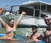 A group of people is enjoying a sunny day in the water near a boat named COASTAL CRUISES with some holding drinks and one woman cheerfully raising her arm in the air