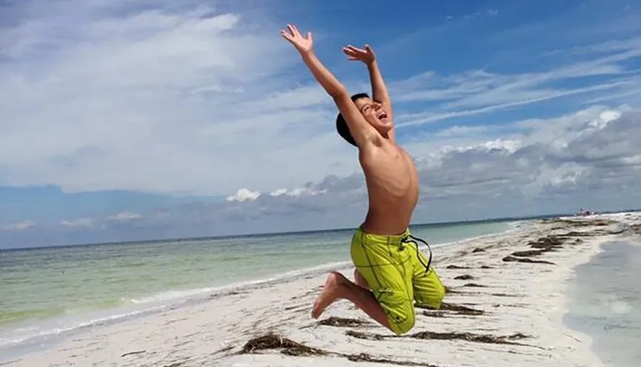 A child is captured in mid-air, joyfully jumping on a sandy beach under a blue sky scattered with clouds.