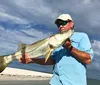 A man in sunglasses and a hat proudly holds a large fish with a beach and cloudy sky in the background