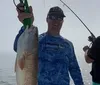 A man in sunglasses and a hat proudly holds a large fish with a beach and cloudy sky in the background