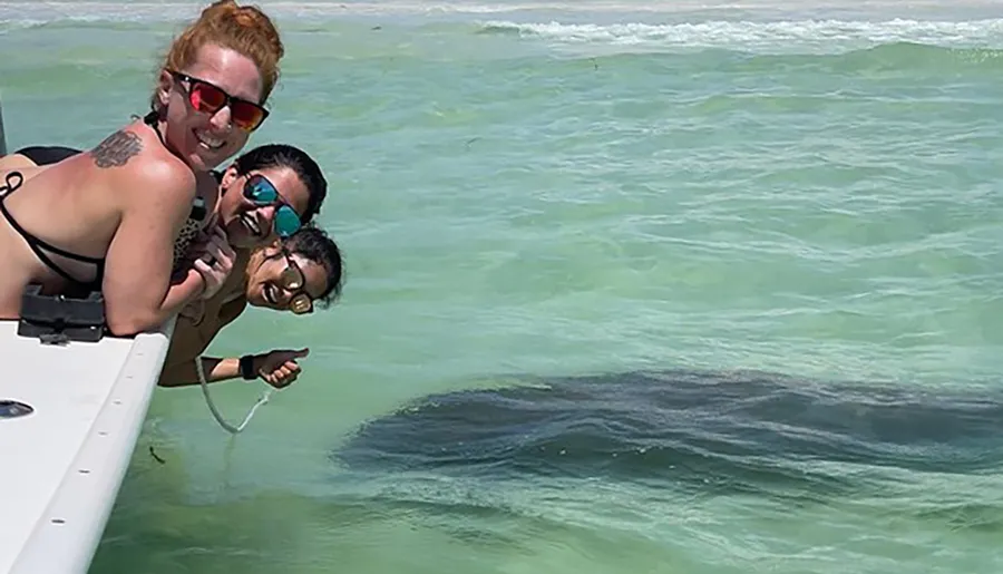 Three people in sunglasses are posing with excitement at the edge of a boat as they observe a large sea creature swimming below the water's surface.