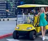 A smiling woman in a green dress is standing next to a yellow golf cart on a city street gesturing with her arm outstretched
