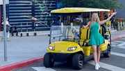 A smiling woman in a green dress is standing next to a yellow golf cart on a city street, gesturing with her arm outstretched.