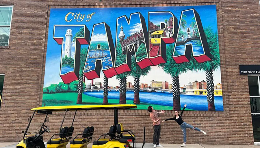 Two people pose in front of a colorful mural that spells out City of TAMPA with scenes of the city depicted within each letter.