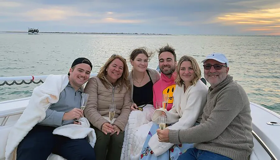 A group of six people is smiling for a photo while enjoying a boat ride together, with glasses in hand and a sunset in the background.