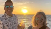 A smiling man and woman are enjoying a sunset on a boat, holding glasses of wine.