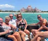 A group of five people are smiling and enjoying a sunny boat ride near a beach with a large pink building in the background