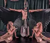 A performer with large outstretched fan-like wings is flanked by two kneeling figures all in theatrical costumes suggesting a scene from a dramatic performance or dance