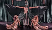 A performer with large, outstretched, fan-like wings is flanked by two kneeling figures, all in theatrical costumes, suggesting a scene from a dramatic performance or dance.