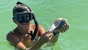 A child wearing a diving mask is holding a conch shell above the water surface.