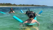 Two people are snorkeling in clear blue water with one person in the foreground wearing a snorkel mask and another slightly in the background.