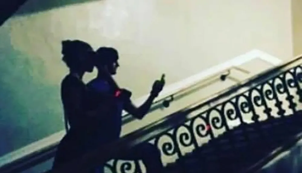 The image depicts the silhouette of two individuals on a staircase one seemingly taking a photo with a smartphone