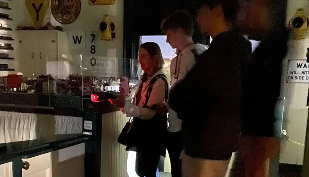 Three people are gathered in a dimly lit room with one person holding a smartphone all of them near a window with various objects and signage visible in the background