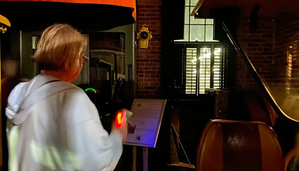 A person with light-colored hair wearing a white hoodie and holding what appears to be a sci-fi styled toy gun is looking at a clipboard illuminated by the guns light in a dimly-lit room with brick walls and a staircase railing visible in the background