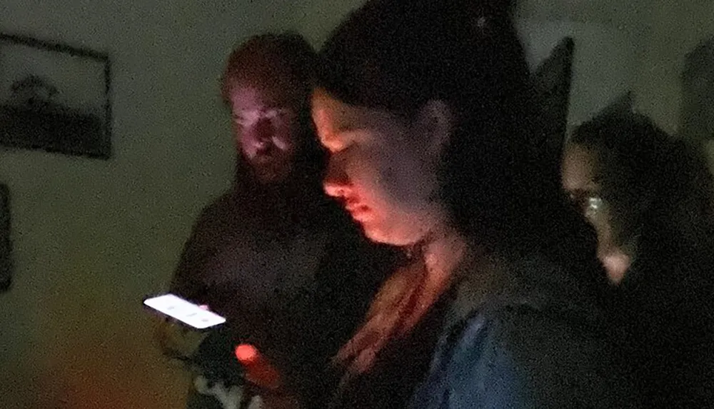 In a dimly lit room a person is looking intently at their smartphone screen illuminating their face while another person appears in the background with a faint glow on their face