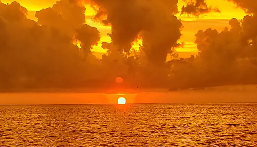 The image captures a vibrant sunset with the sun dipping into the ocean horizon, framed by dramatic orange-tinted clouds.