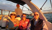 Three people are smiling for the camera while enjoying a boat ride at sunset, with another person facing away toward the water.