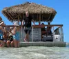 A group of people enjoys a sunny day on a floating dock with a thatched roof in shallow waters