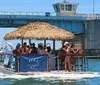 A group of people enjoys a sunny day on a floating dock with a thatched roof in shallow waters