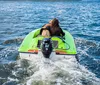 Two individuals are riding a green mini powerboat in a marina with other boats and yachts docked in the background