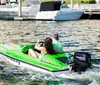 Two individuals are riding a green mini powerboat in a marina with other boats and yachts docked in the background