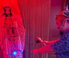 A person wearing a headband and sunglasses is using a device to interact with a figure draped in cloth under red lighting creating a spooky atmosphere