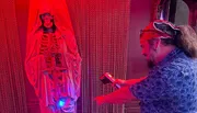 A person wearing a headband and sunglasses is using a device to interact with a figure draped in cloth under red lighting, creating a spooky atmosphere.