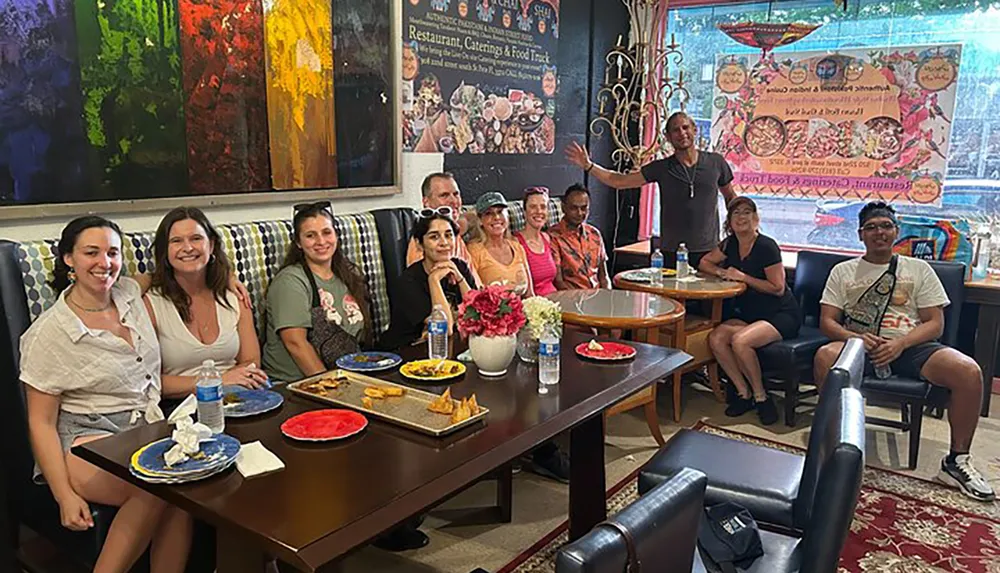 A group of people are smiling and posing for a photo in a colorful restaurant with artwork on the walls and food on their tables