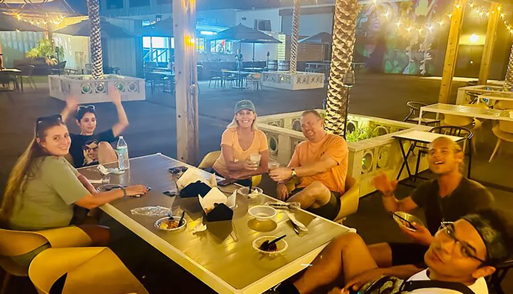 A group of people are enjoying a lively evening at an outdoor dining area with festive lights and street art in the background