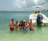 A group of people is enjoying a sunny day in shallow waters beside a boat with some holding beverages and the coastline visible in the background