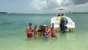 A group of people is enjoying a sunny day in shallow waters beside a boat, with some holding beverages and the coastline visible in the background.