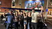 A group of six smiling people pose in front of a colorful mural that says 
