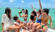 A group of people are enjoying a sunny day on a boat, with one raising her arm in excitement and another drinking from a bottle, amidst a backdrop of clear turquoise waters and other boats.