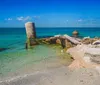 The image shows a bright blue seascape with an old dilapidated pier or structure made of concrete and stone partially submerged in the clear waters near a sandy beach