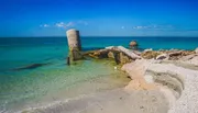 The image shows a bright blue seascape with an old, dilapidated pier or structure made of concrete and stone, partially submerged in the clear waters near a sandy beach.