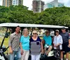 A group of seven people are smiling for a photo in front of a white open-air shuttle with trees and a cityscape in the background