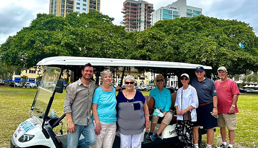 A group of seven people are smiling for a photo in front of a white open-air shuttle with trees and a cityscape in the background