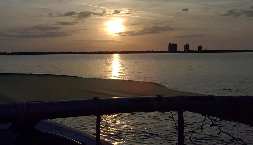 The image captures a serene sunset over a body of water with the suns reflection on the surface seen from the perspective of a boat with a hint of silhouetted infrastructure on the distant shoreline