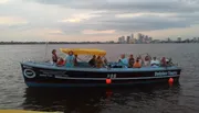 A group of people are enjoying a boat tour at dusk with a city skyline in the background.