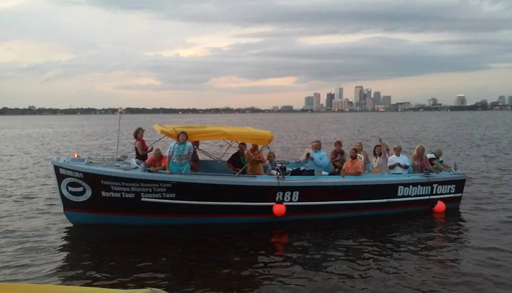 A group of people are enjoying a boat tour at dusk with a city skyline in the background