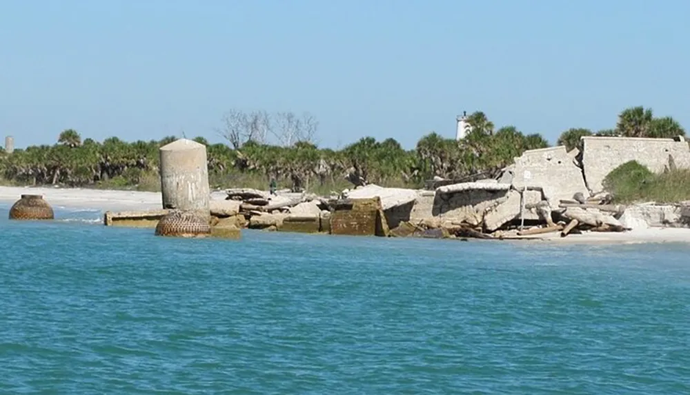 This image shows the ruins of a coastal structure partially submerged and eroded with the ocean in the foreground and a sandy beach with vegetation in the background