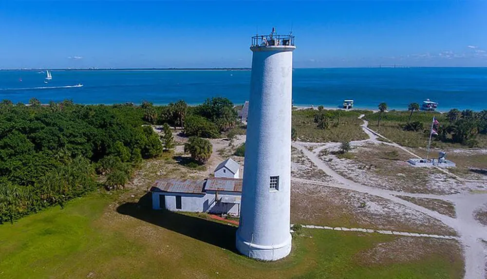 An aerial view of a coastal landscape featuring a tall white lighthouse with surrounding buildings greenery and a blue sea with boats visible in the background