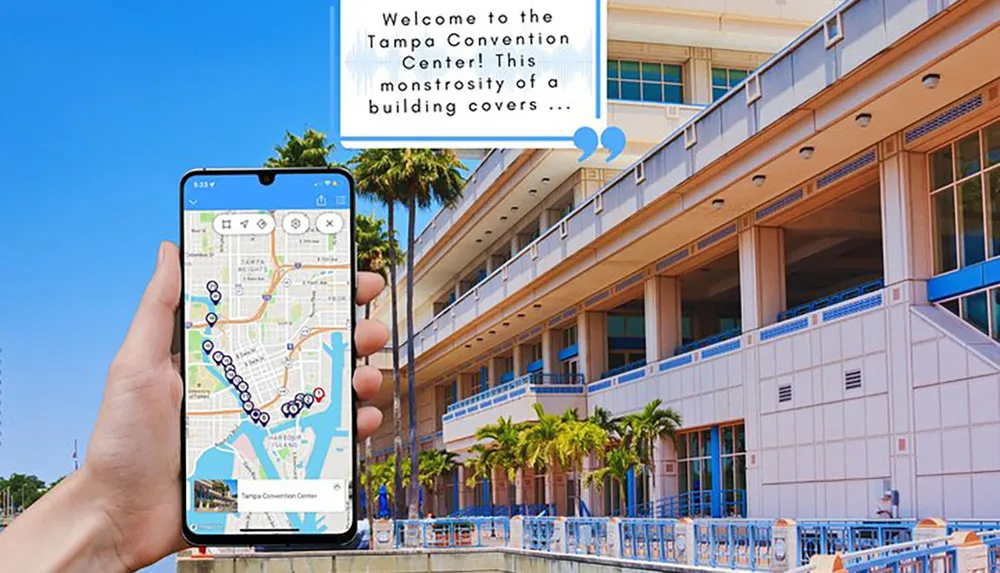 A persons hand is holding a smartphone with a map on the screen displaying the location of the Tampa Convention Center which is shown in the background of the photo