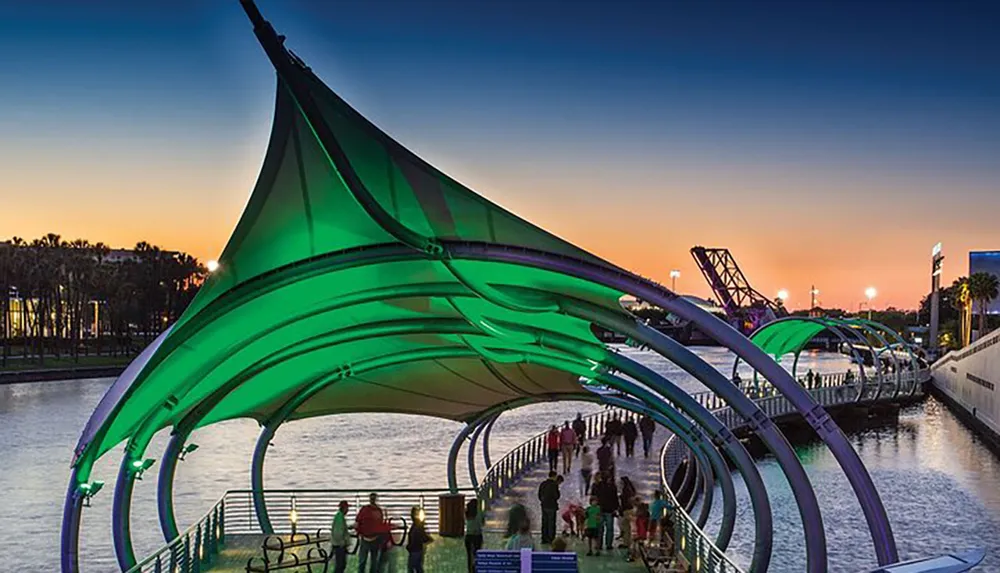 The image shows a lively waterfront walkway with a modern illuminated green canopy at dusk with people gathered along its length