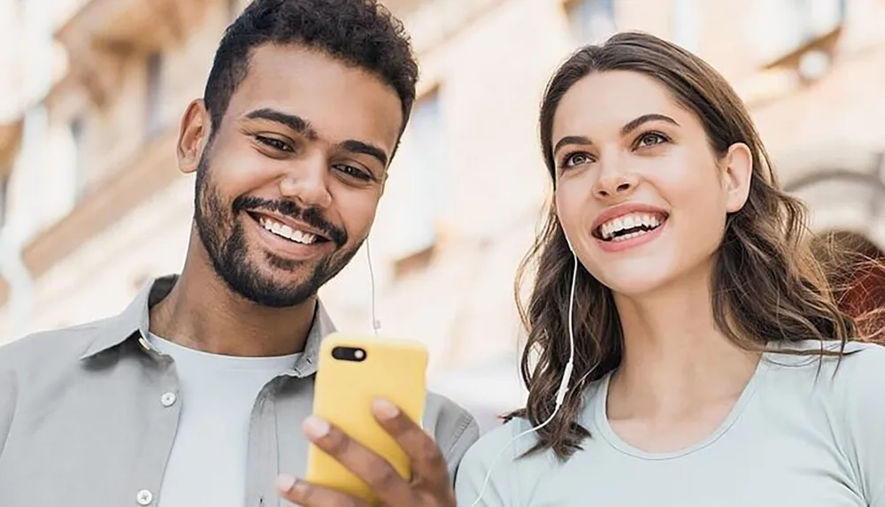 A smiling man and woman are sharing earbuds and looking at a smartphone that the man is holding