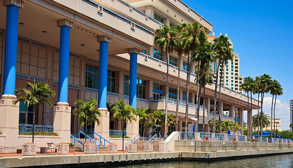 The image showcases a modern waterfront building with blue columns flanked by palm trees against a background of a blue sky