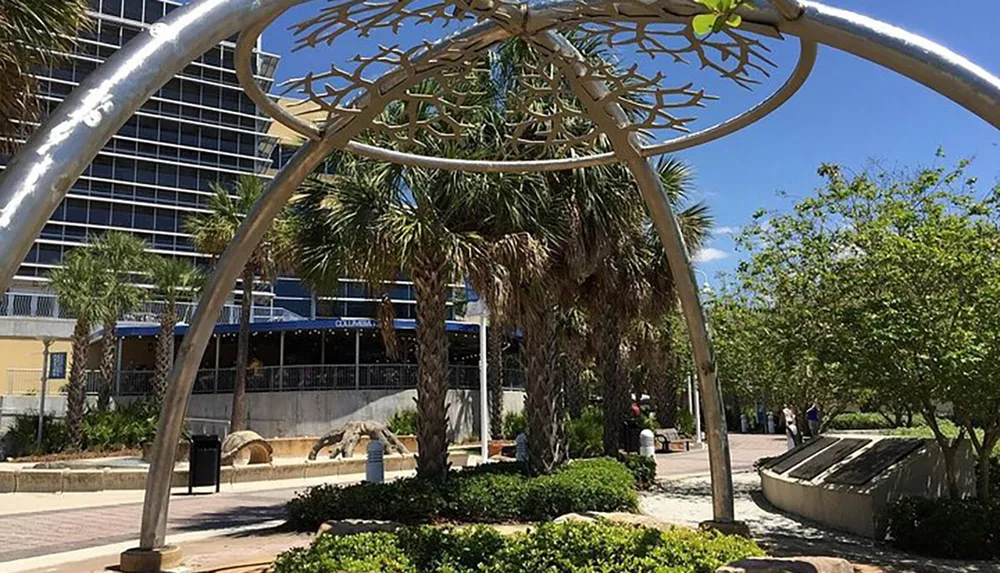 The image shows a sunny outdoor scene with a unique metallic tree-like sculpture in the foreground palm trees and a building with shaded seating areas in the background