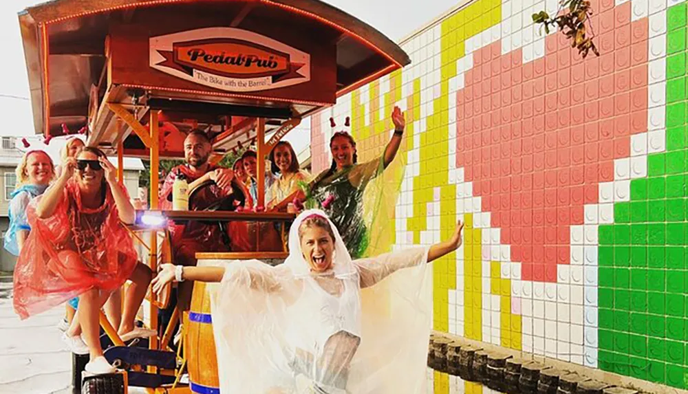 A group of people wearing ponchos is enjoying a ride on a pedal-powered pub on wheels with one exuberant individual in front extending their arms widely