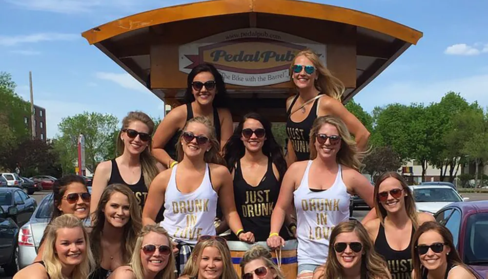 A group of cheerful people are enjoying a ride on a Pedal Pub with some wearing thematic tank tops with playful phrases about being drunk or in love