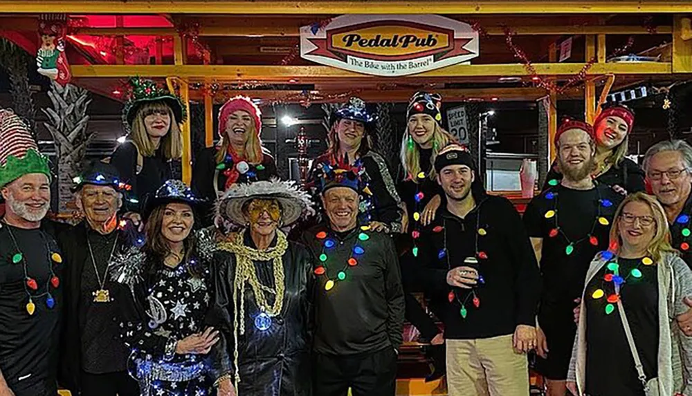 A group of merry individuals in festive attire and holiday lights poses for a photo on a Pedal Pub suggesting a celebration or party on wheels